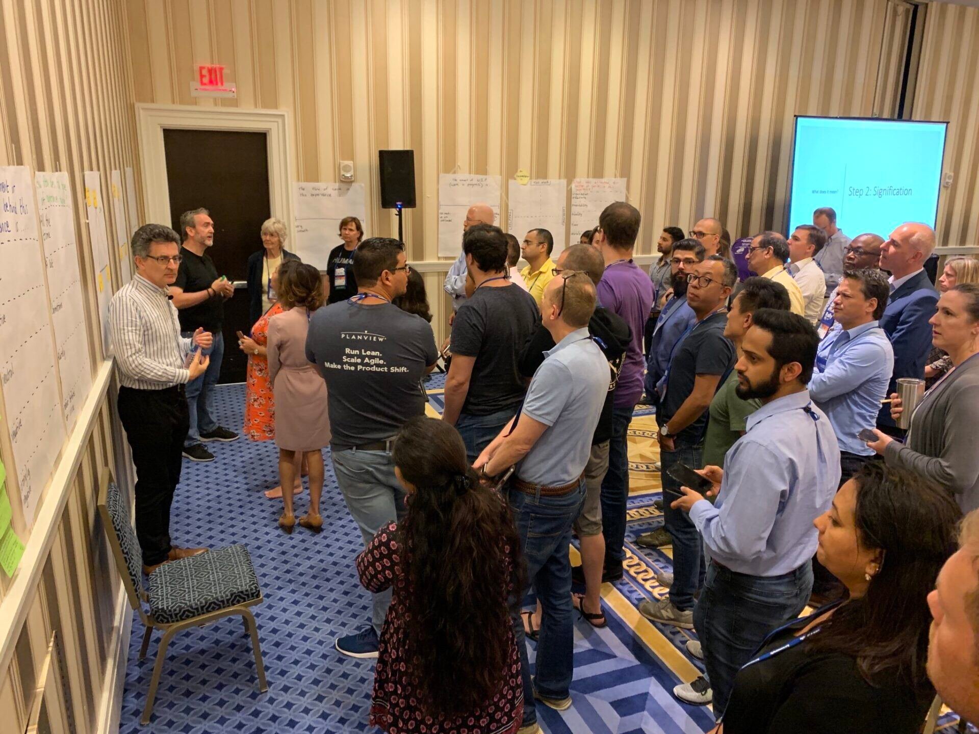 A facilitator leads a large group of professionals through a collaborative strategy workshop, with wall-mounted notes and a presentation screen visible in the background.