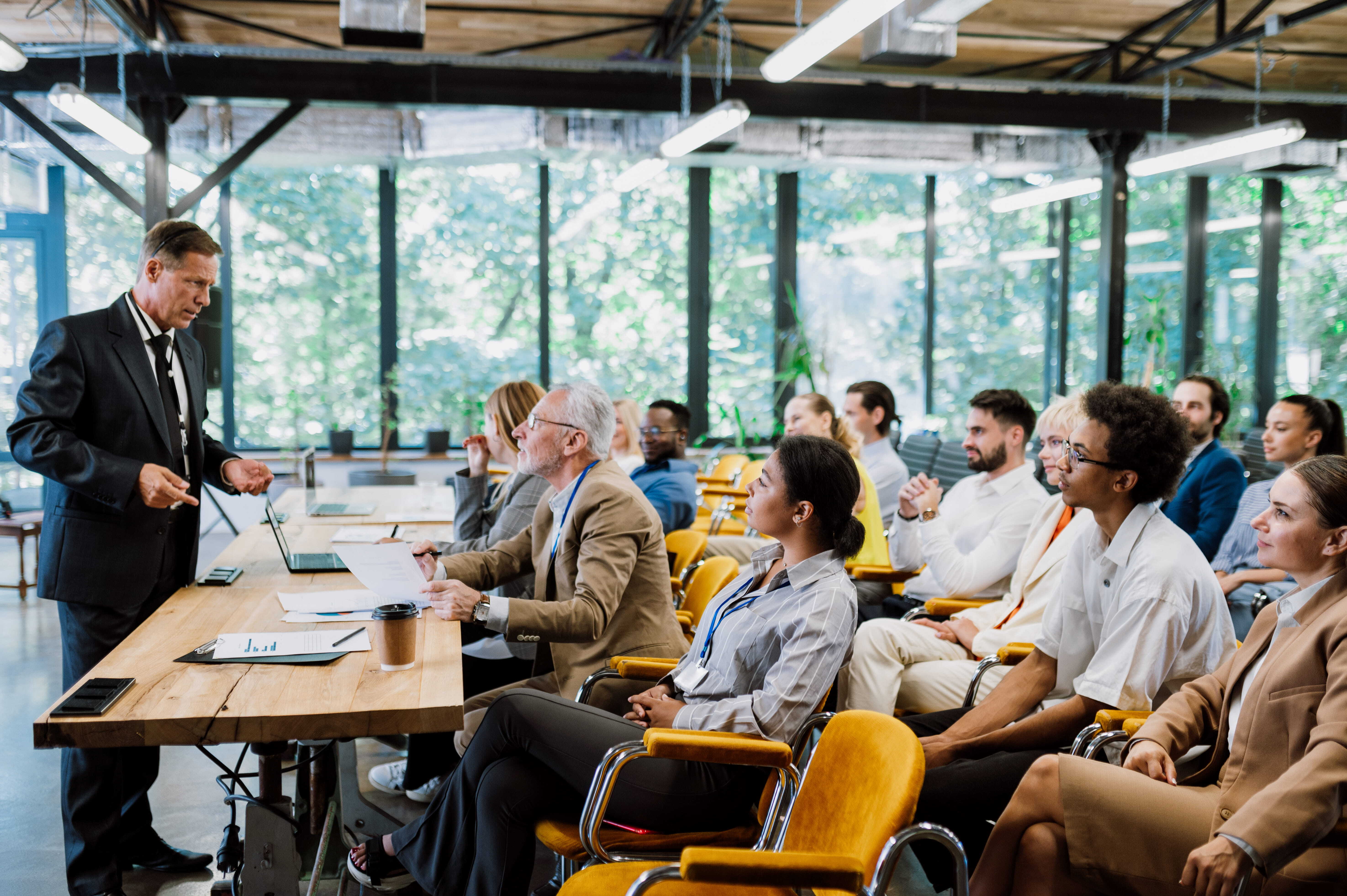 A senior business consultant presents to a group of engaged professionals in a modern, light-filled meeting room, illustrating the dynamic of expert advisory and strategic dialogue.