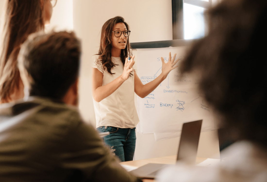 A woman speaks to colleagues during a workshop, using practical examples on a flip chart to explain ideas and guide a leadership discussion.