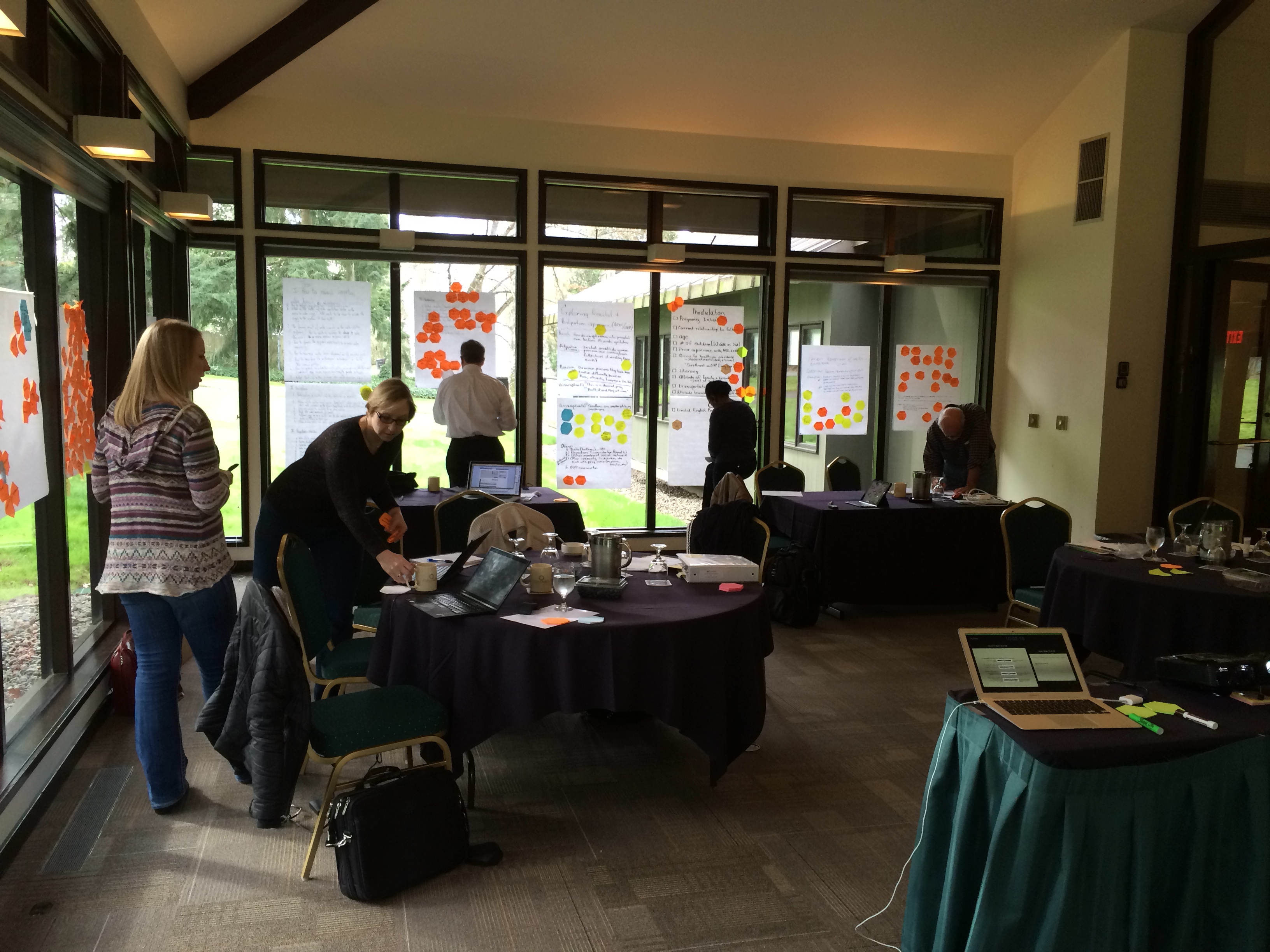 Workshop participants collaborate at round tables with laptops and colourful sticky notes on windows during complexity training session.
